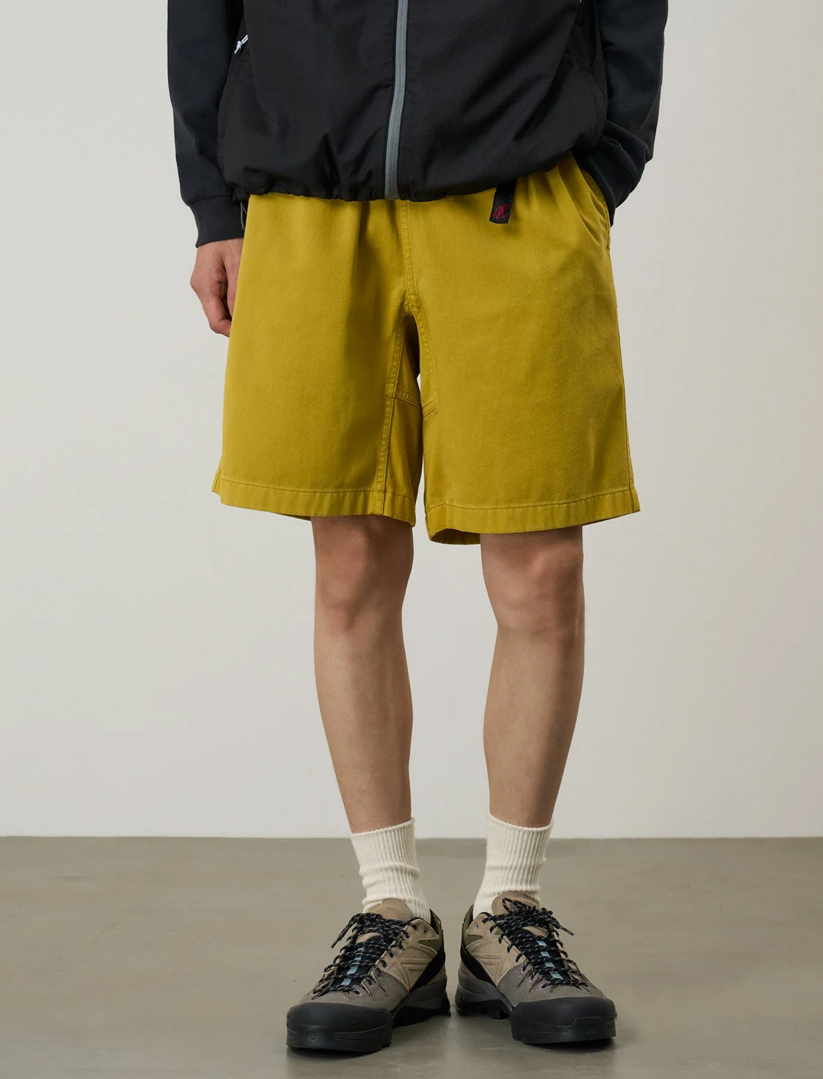 Person wearing gramicci g-short mustard yellow shorts and a black jacket on a plain background