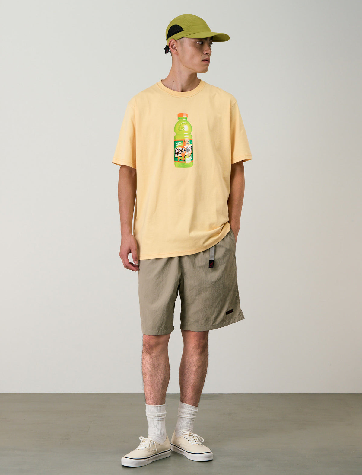 Person wearing a yellow t-shirt with a bottle graphic, beige shorts, and a green cap on a plain background