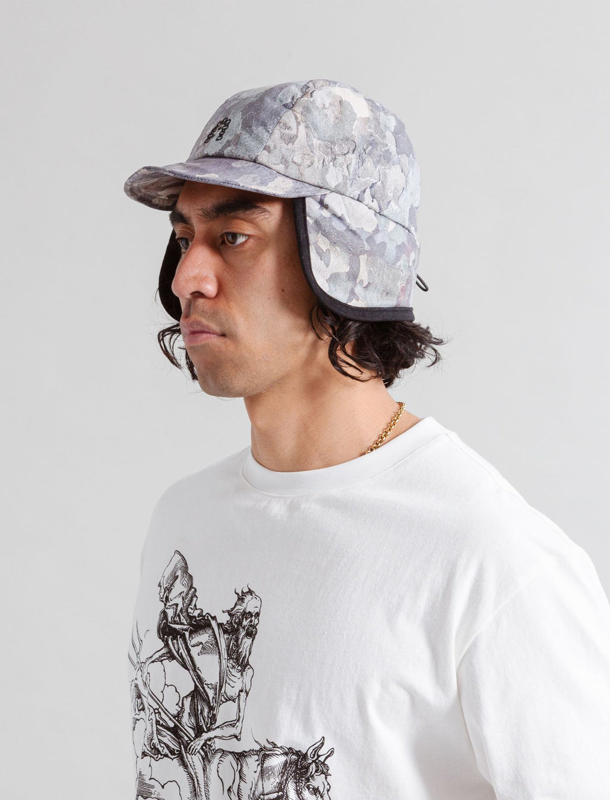 Person wearing a camouflage cap and white t-shirt with graphic design on a gray background