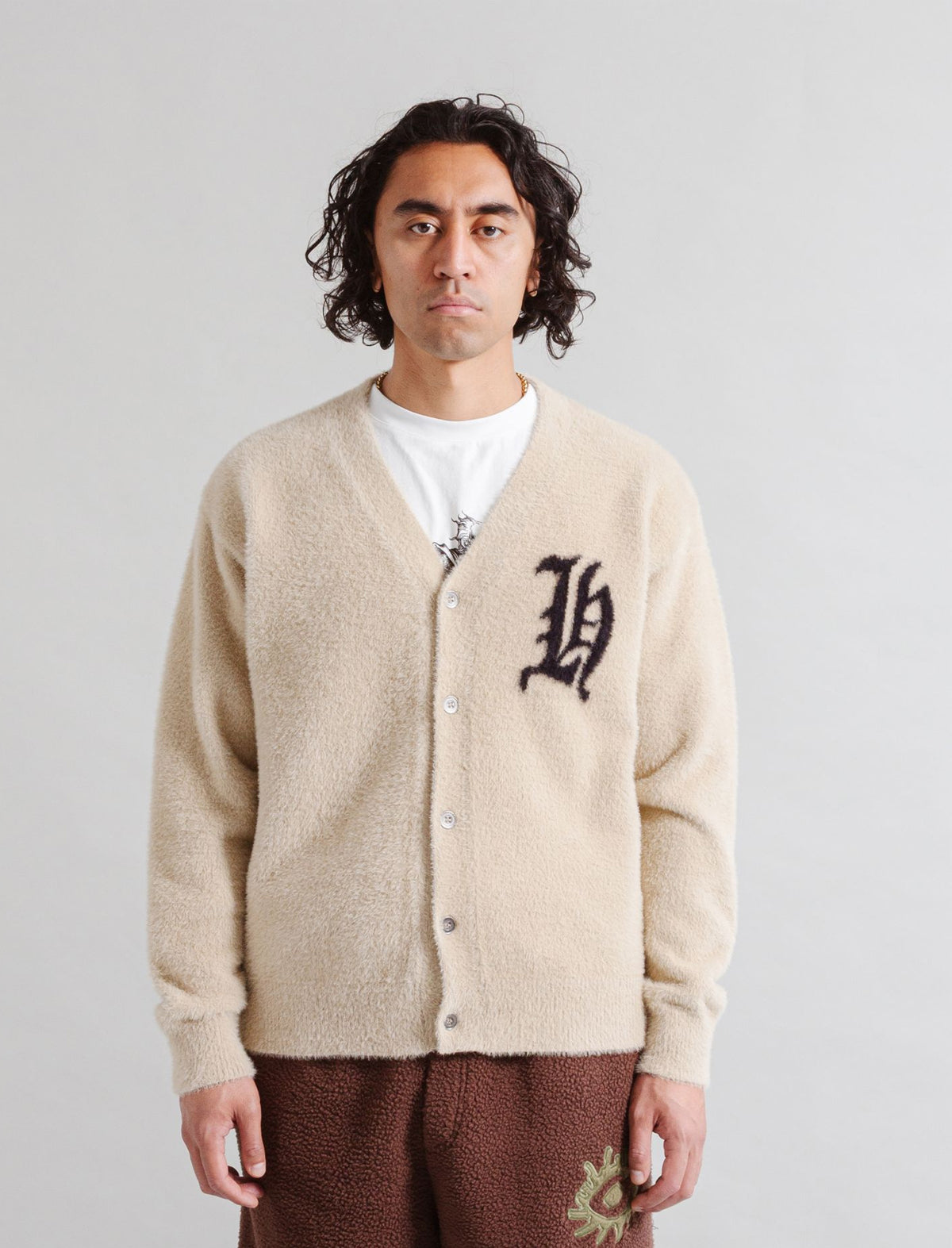 Person wearing a Heresy 'H' beige cardigan with a logo, standing against a plain background