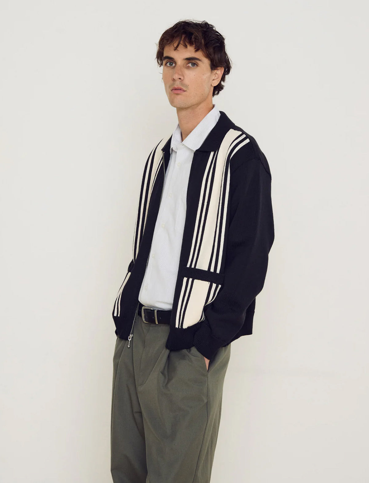 Man wearing a black and white striped sweater over a white shirt with gray pants on a plain background