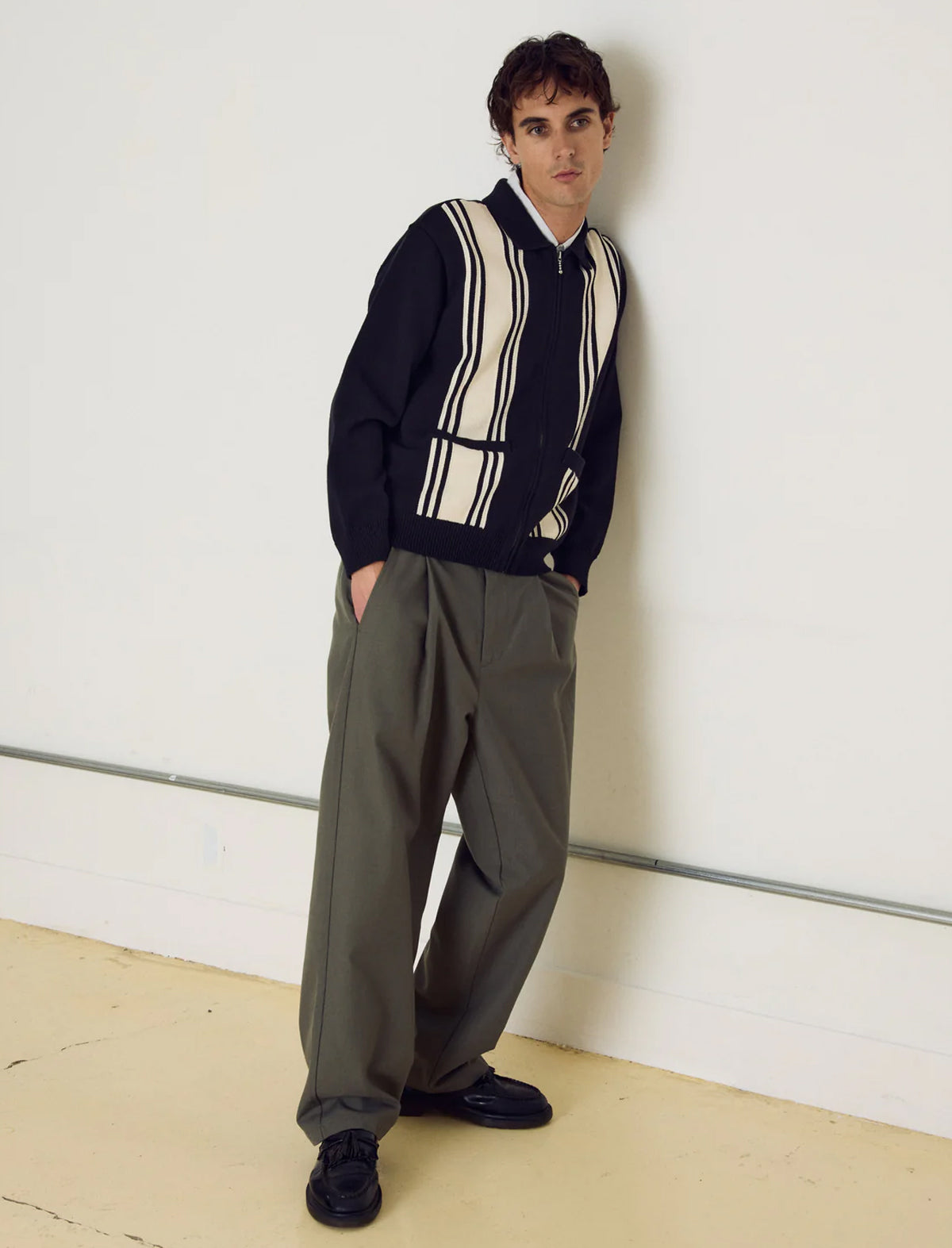 Man wearing a dark sweater with a striped pattern and light-colored pants against a white wall.