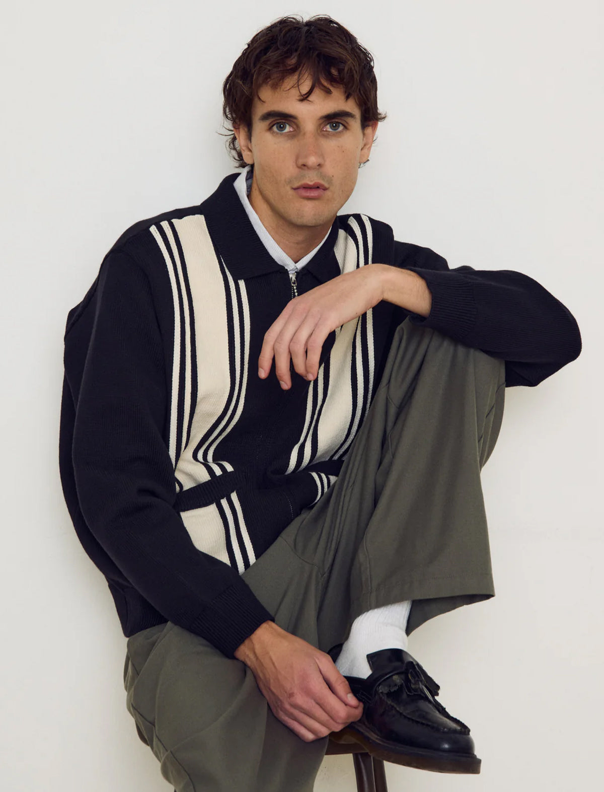 Man wearing a navy sweater with white stripes and gray pants sitting on a stool against a white wall.