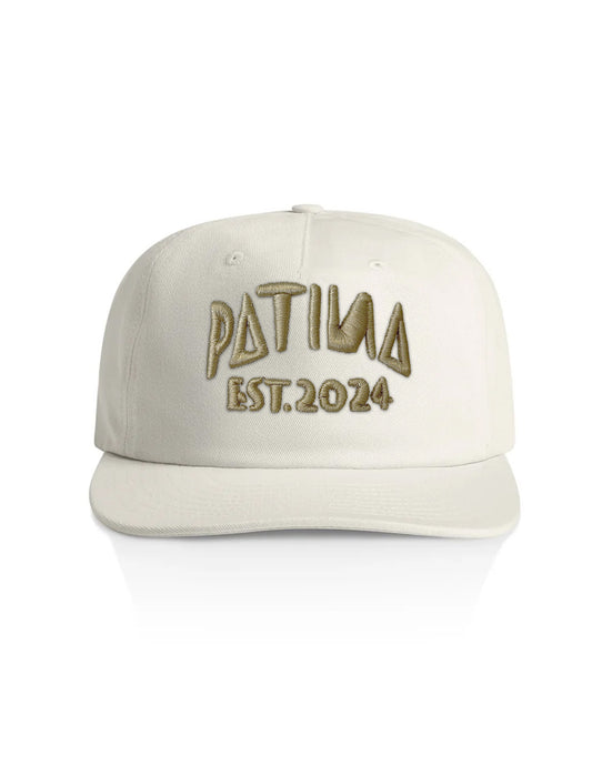 An off-white structured baseball cap with 'PATINA EST. 2024' embroidered on the front in a metallic gold tone, set against a plain white background.