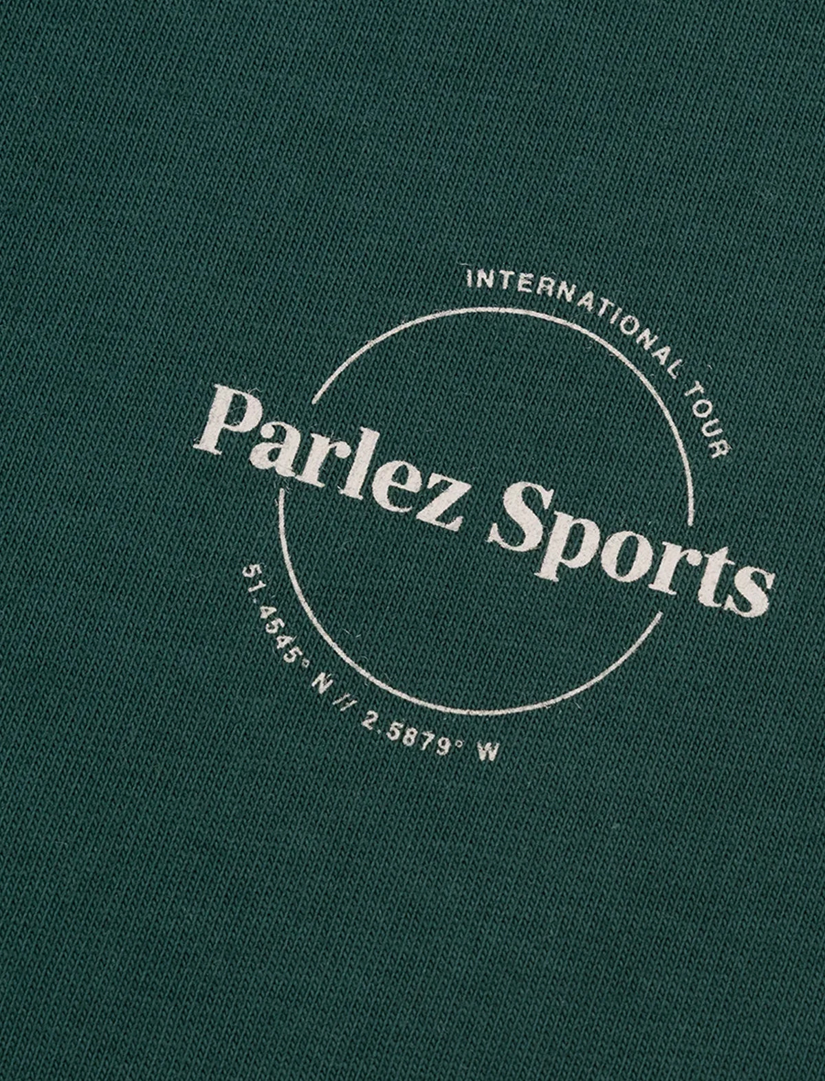 Close-up of a dark green textured fabric with a white circular logo reading 'Parlez Sports' and smaller text 'International Tour' and coordinates.