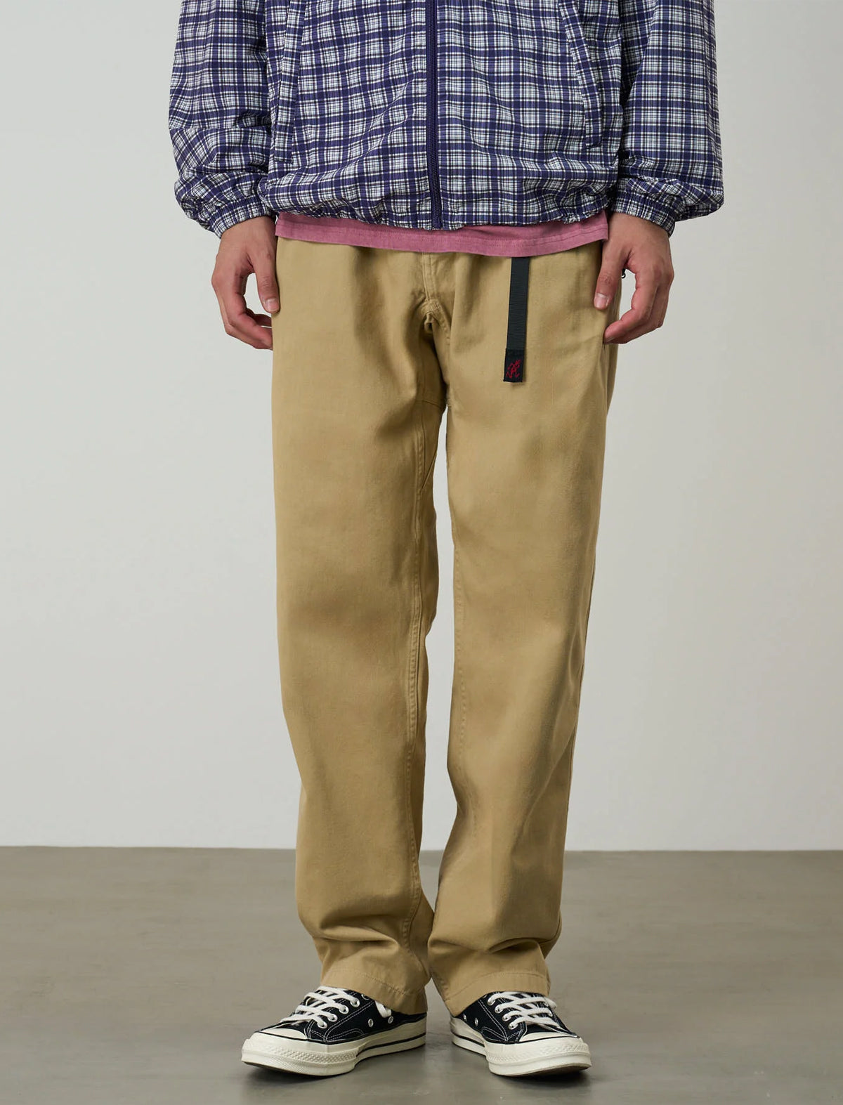 A person wearing tan straight-leg work pants, a blue and white plaid zip-up jacket, and black canvas sneakers, standing against a plain light gray studio background.