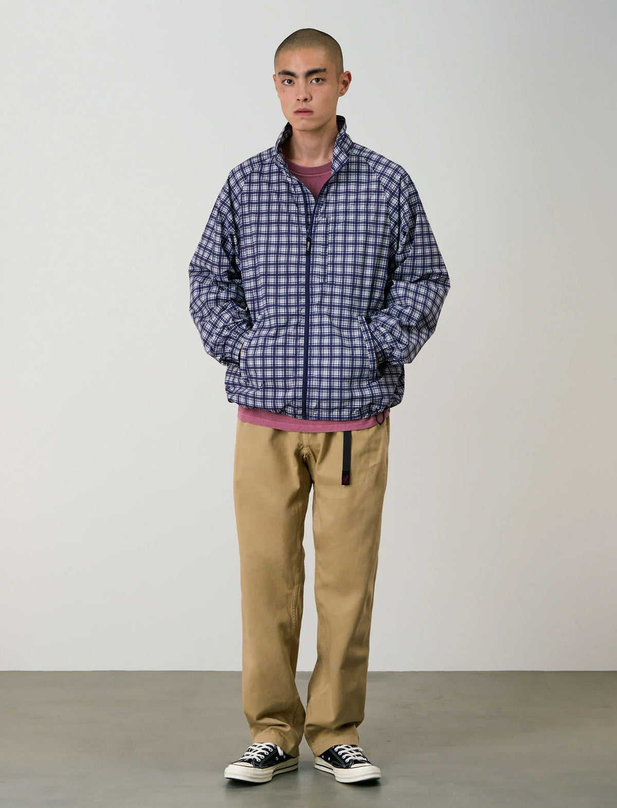 A man stands against a plain light gray studio background wearing a blue and white checkered zip-up jacket over a mauve shirt, tan pants, and black-and-white sneakers.