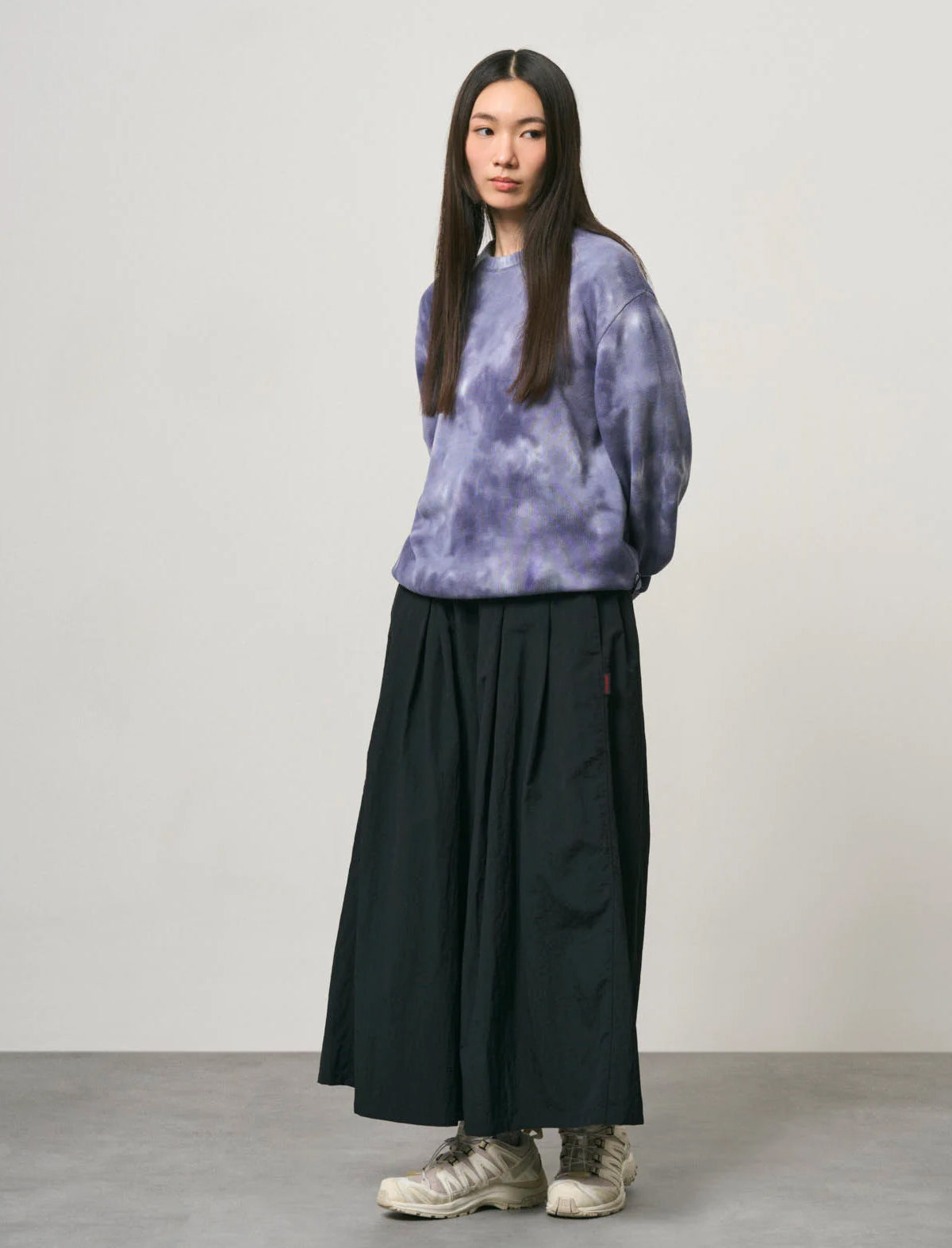 A woman stands in a studio wearing a purple and blue tie-dye sweatshirt, a long black pleated skirt, and beige sneakers against a light gray background.