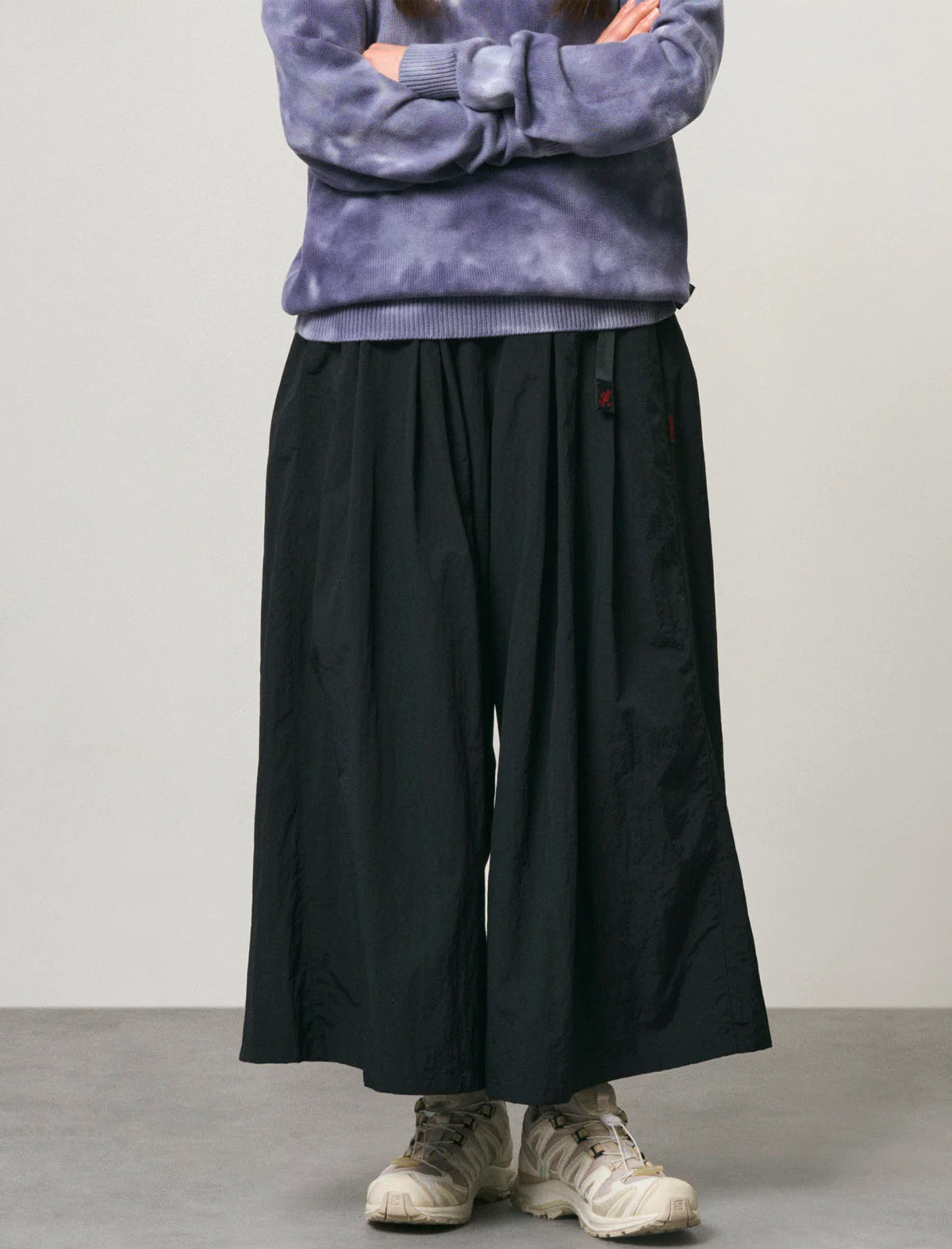 A person wearing loose black wide-leg pants, a purple tie-dye sweatshirt, and off-white sneakers, standing against a plain light gray studio background.