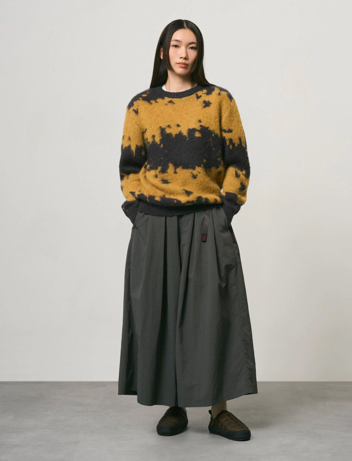 A model stands against a plain light gray studio wall wearing a mustard yellow and black tie-dye knit sweater paired with a dark charcoal gray pleated skirt and dark slip-on shoes.