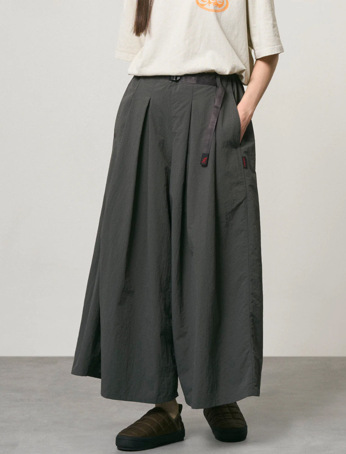 A person wearing dark charcoal gray wide-leg pleated pants with a drawstring waist, paired with a cream t-shirt and dark brown shoes, standing against a neutral light gray studio background.