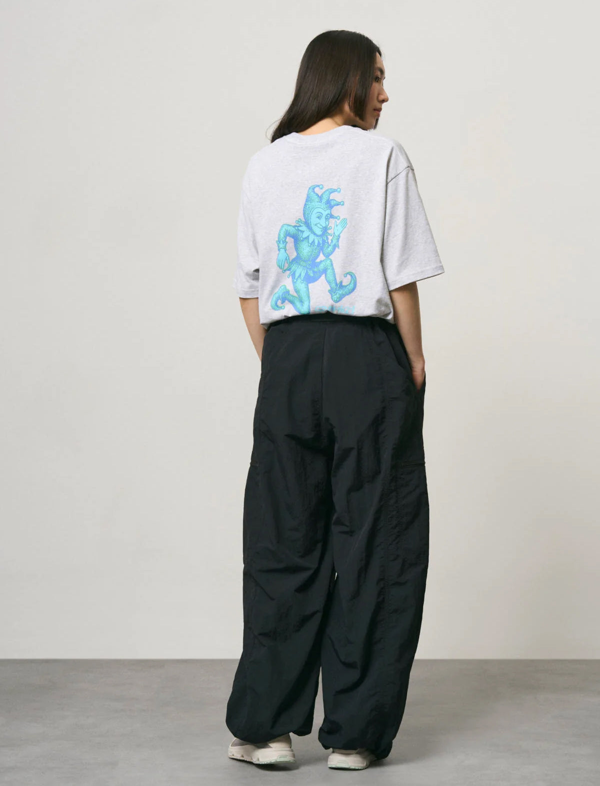 A model viewed from behind wearing a light gray oversized t-shirt with a large blue dragon graphic on the back, paired with black wide-leg cargo pants and white sneakers, set against a minimalist studio background.