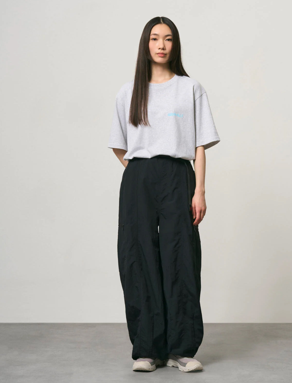 A woman stands in a studio wearing an oversized light gray T-shirt with a small light blue chest graphic, loose black wide-leg pants, and light-colored sneakers.