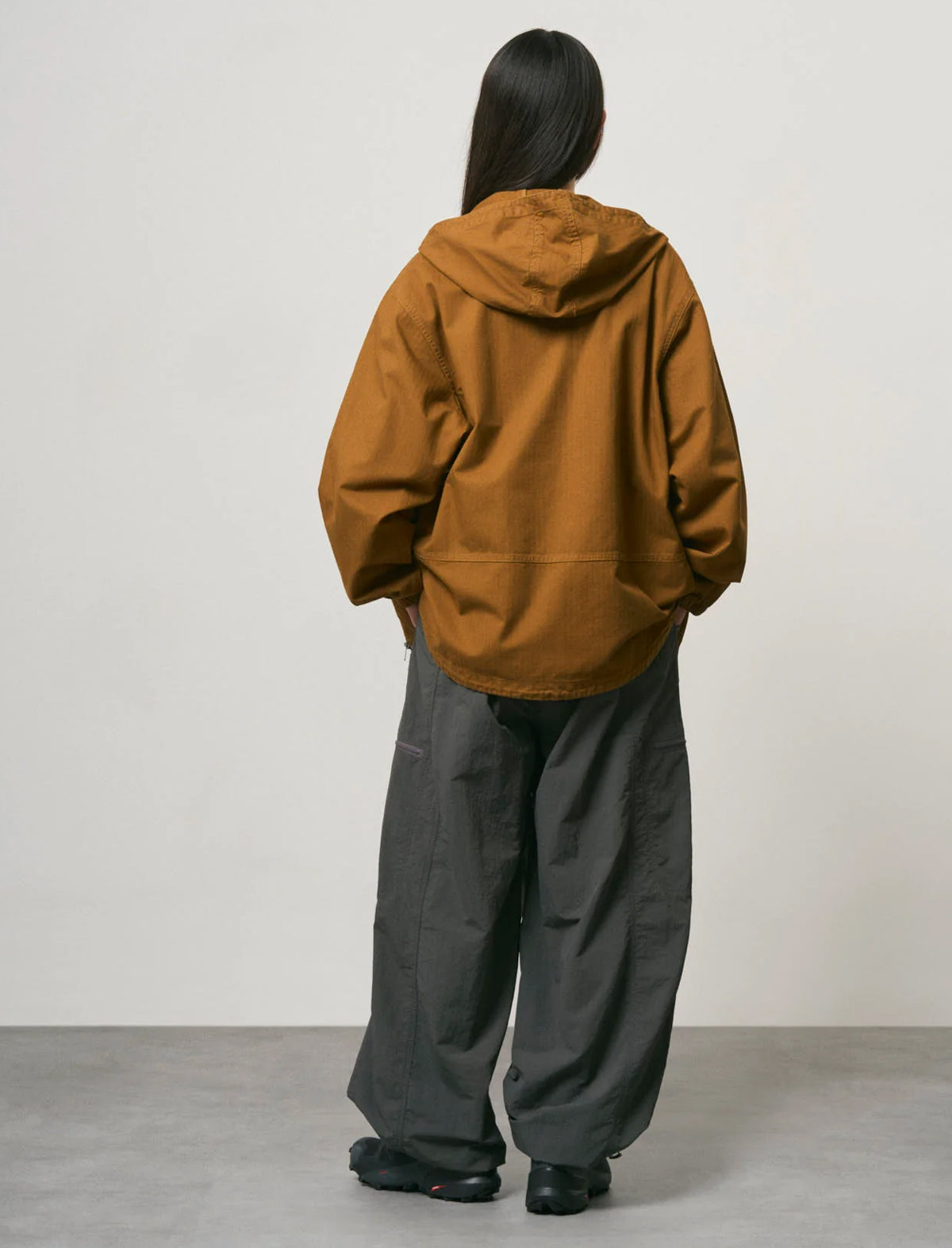 A person shown from the back wearing an oversized mustard-brown hooded jacket and loose dark gray pants, standing against a plain light gray wall and floor.