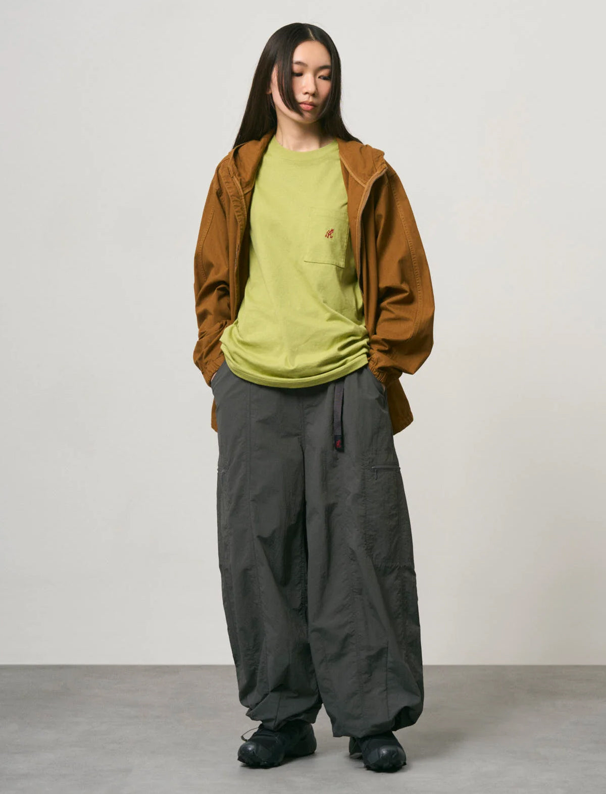 A model stands against a plain light gray studio wall wearing a brown hooded jacket over a lime green t-shirt, paired with loose dark gray cargo pants and black shoes.