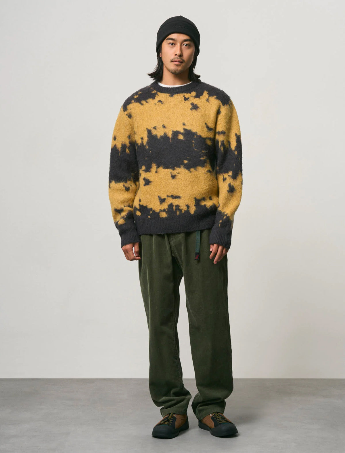 A model stands against a light gray studio background wearing a yellow and black tie-dye knit sweater, dark olive green pants, a black beanie, and dark brown shoes.