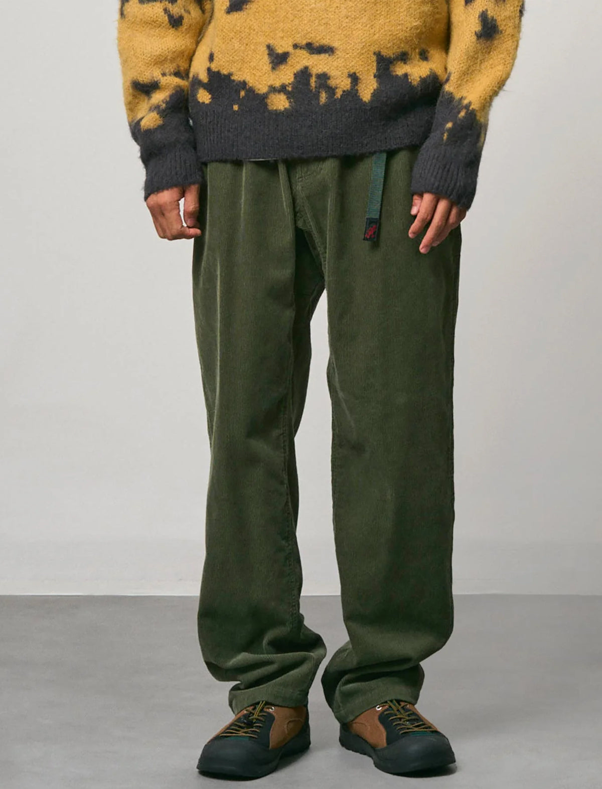 A person shown from the chest down wearing olive green relaxed-fit pants, a yellow and dark gray tie-dye sweater, and brown and black sneakers against a plain studio background.