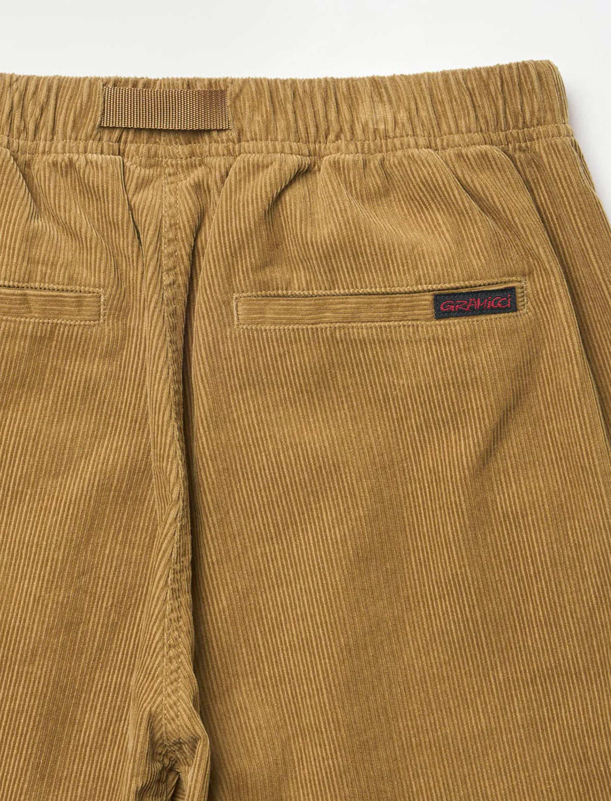 Close-up of a pair of corduroy pants in a warm camel brown color, showing the elastic waistband, back pocket, and a small dark brand label on the side.