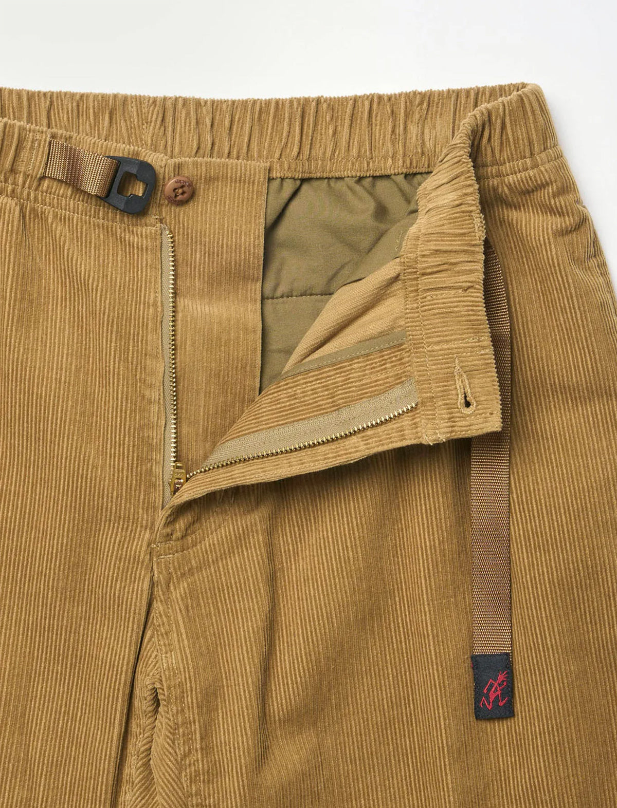 Close-up of tan corduroy trousers showing the waistband, zipper, interior lining, and a small pink embroidered logo patch on the belt loop.
