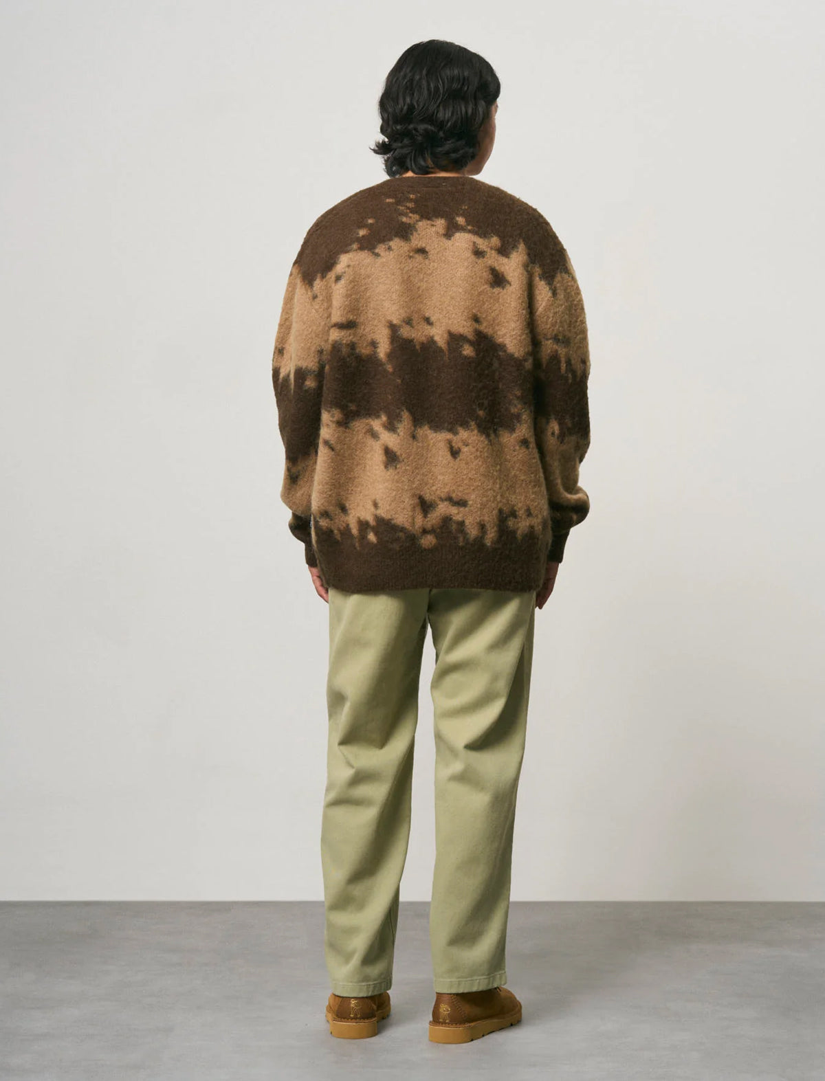 A person standing with their back to the camera, wearing a brown and tan tie-dye knit sweater, olive green pants, and brown suede boots, set against a plain light gray studio background.