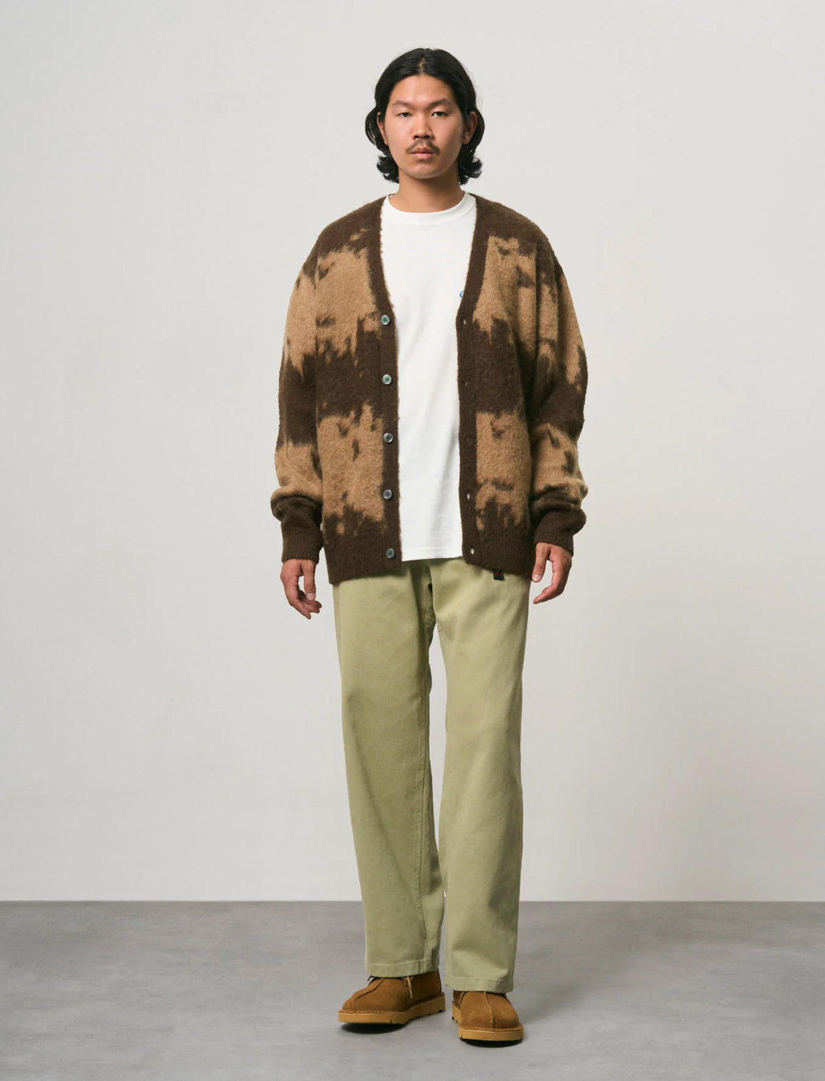 A man stands in a studio wearing a brown and tan patterned knit cardigan over a white crewneck shirt, paired with light olive green pants and brown suede shoes.