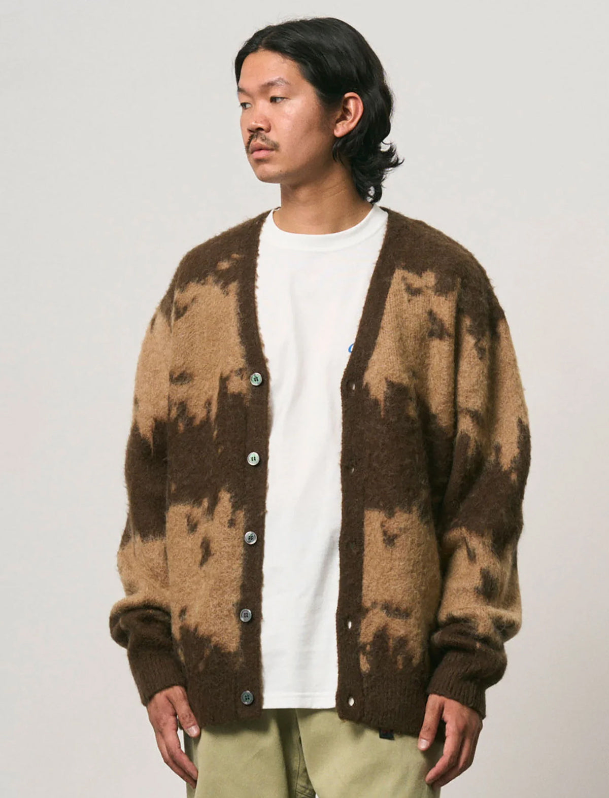 A man wearing a brown and beige tie-dye knit cardigan over a white t-shirt and light olive pants, photographed against a neutral light gray background.