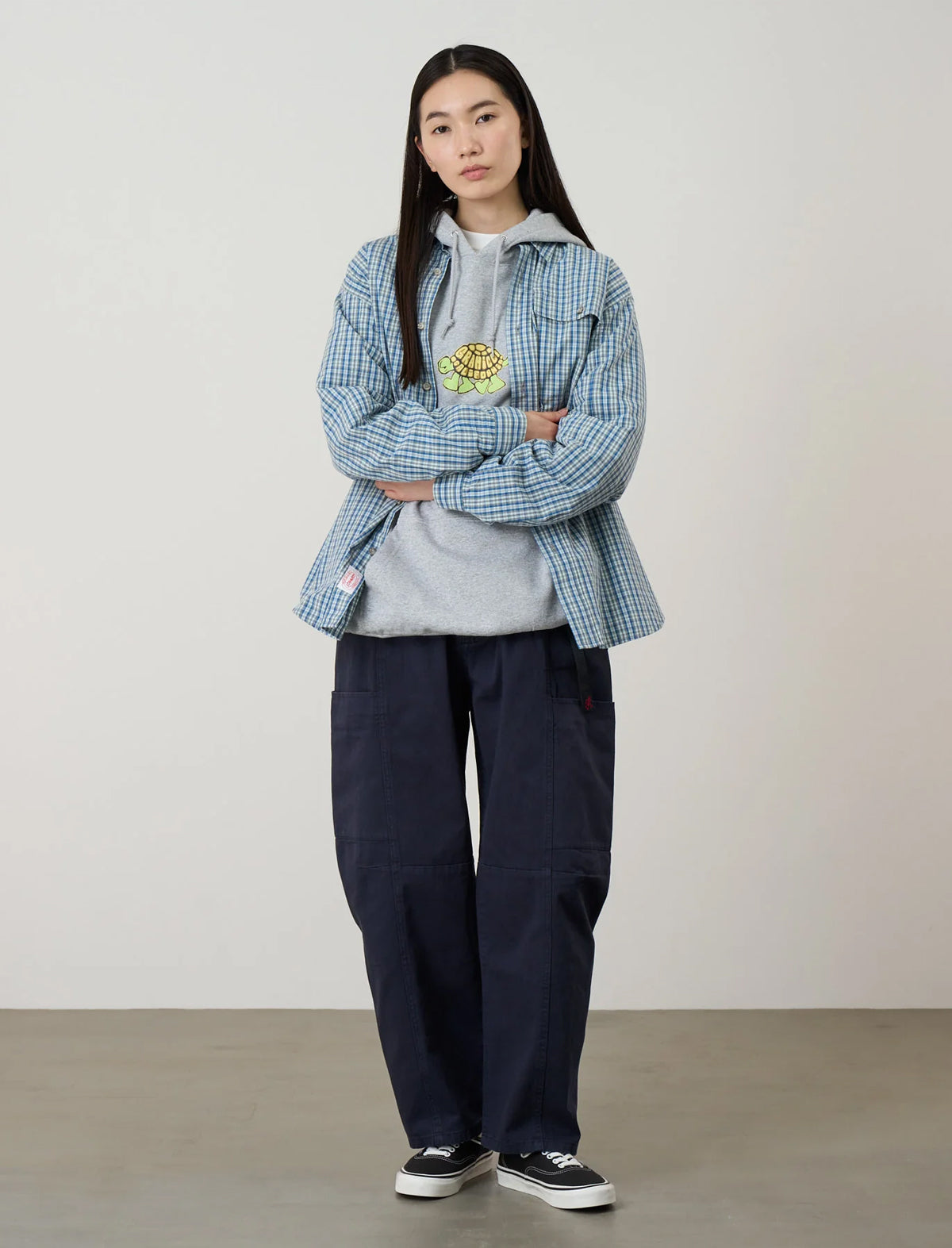 A model stands with arms crossed wearing a light blue and white checkered overshirt with a yellow and green embroidered chest graphic, layered over a light gray hoodie, dark navy wide-leg cargo pants, and black canvas sneakers.