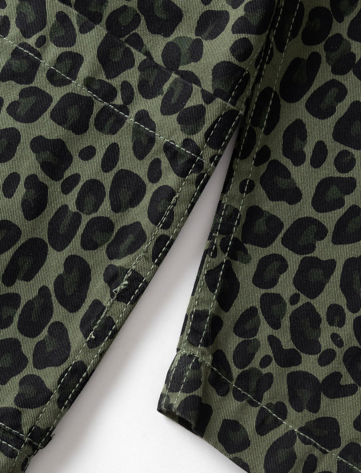 Close-up of a leopard print fabric with an olive green base and black spots, showing stitched seams and a fold over a white background.