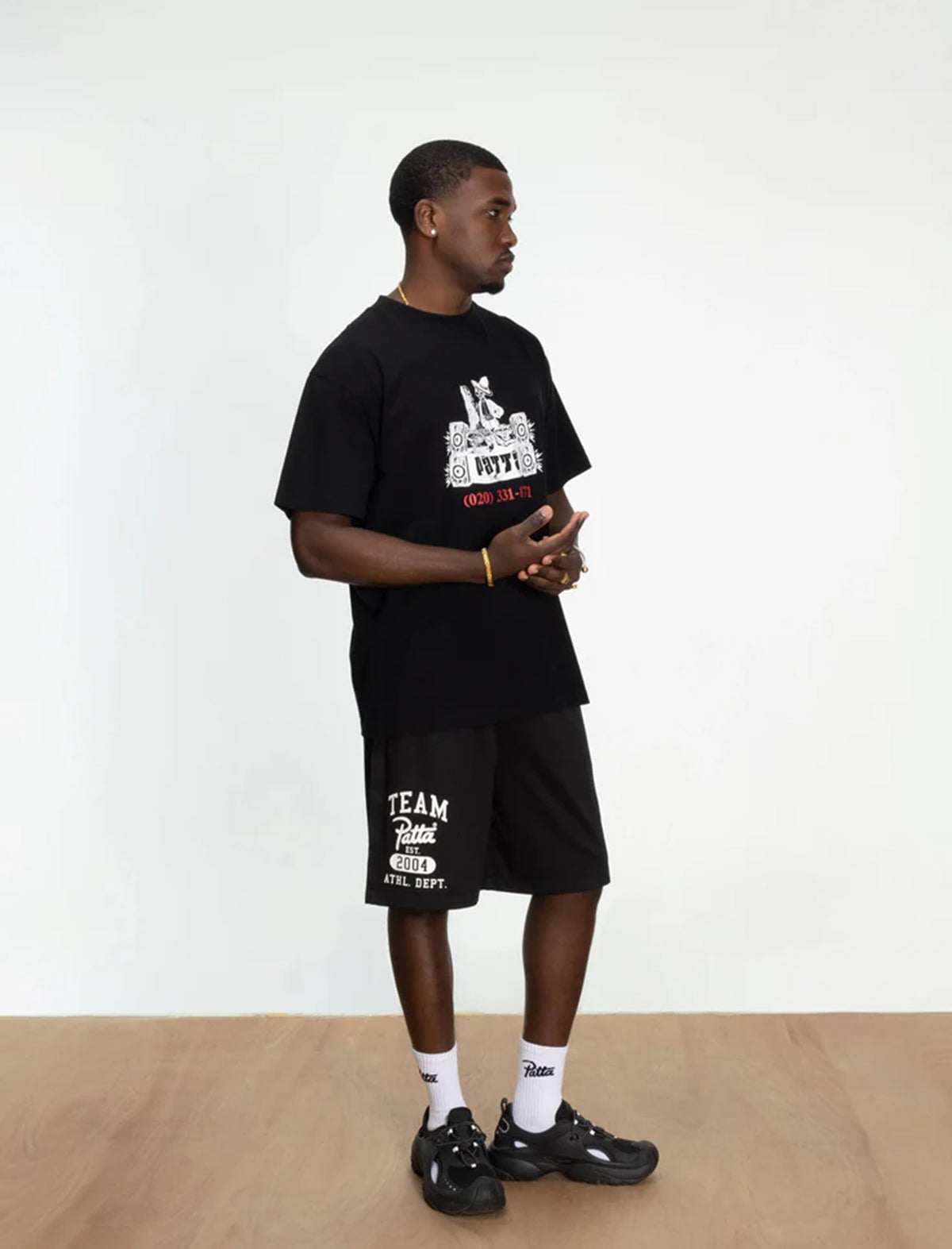 Man wearing a patta black t-shirt and shorts with text, standing against a white wall.