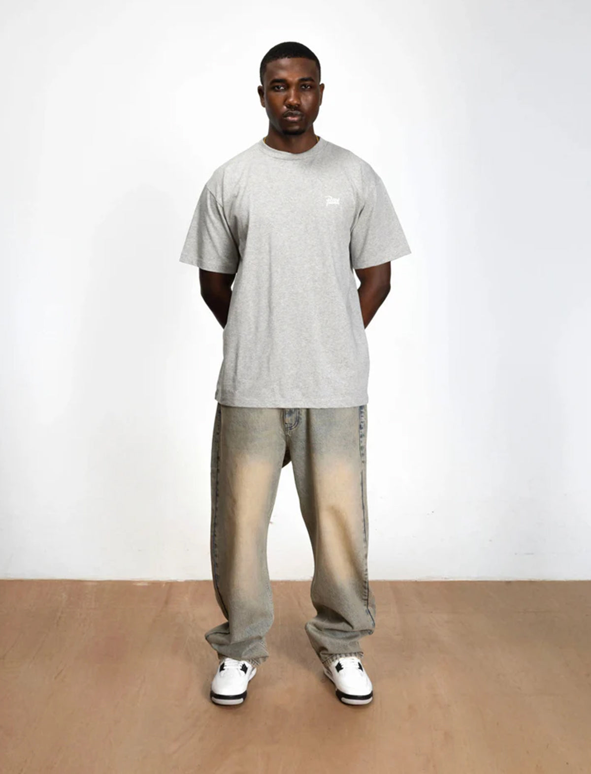 Man wearing a gray t-shirt and light-colored pants standing on a wooden floor with a white background