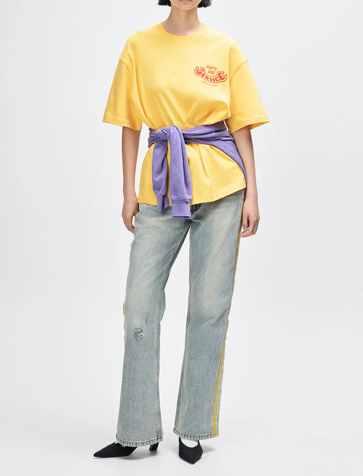 Person wearing a yellow t-shirt with a graphic, light blue jeans, and black shoes on a white background