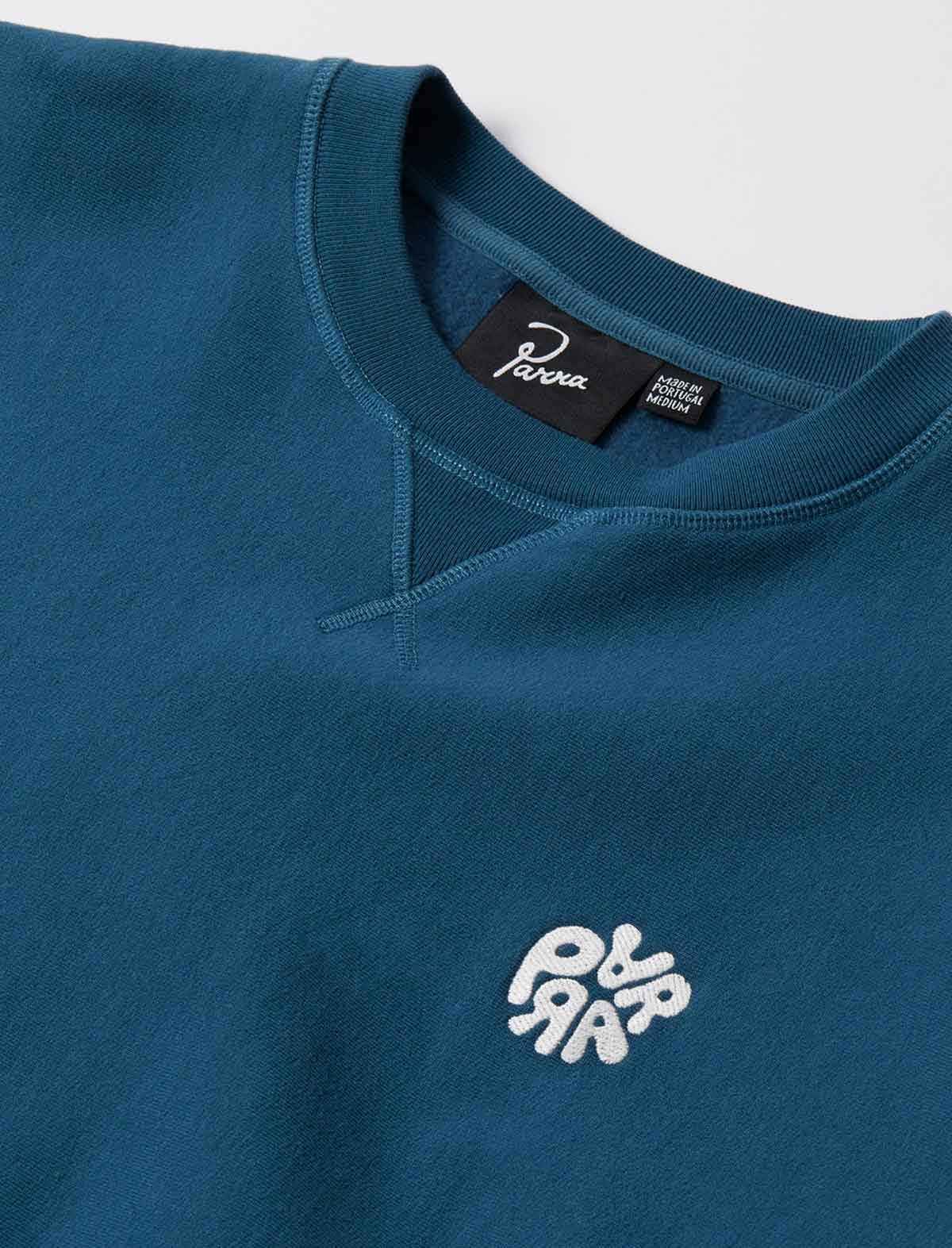 Close-up of a teal crewneck sweatshirt with a white embroidered 'Parra' logo on the chest and a black neck label.