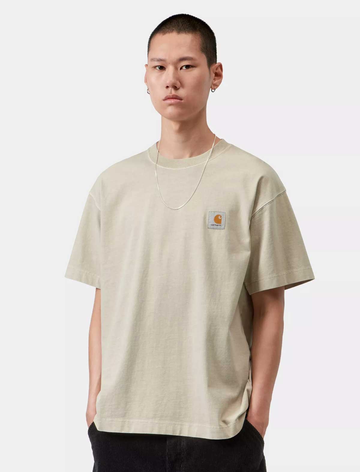 A model wearing a light beige Carhartt t-shirt with the signature chest logo patch, paired with black pants, against a plain light gray background.