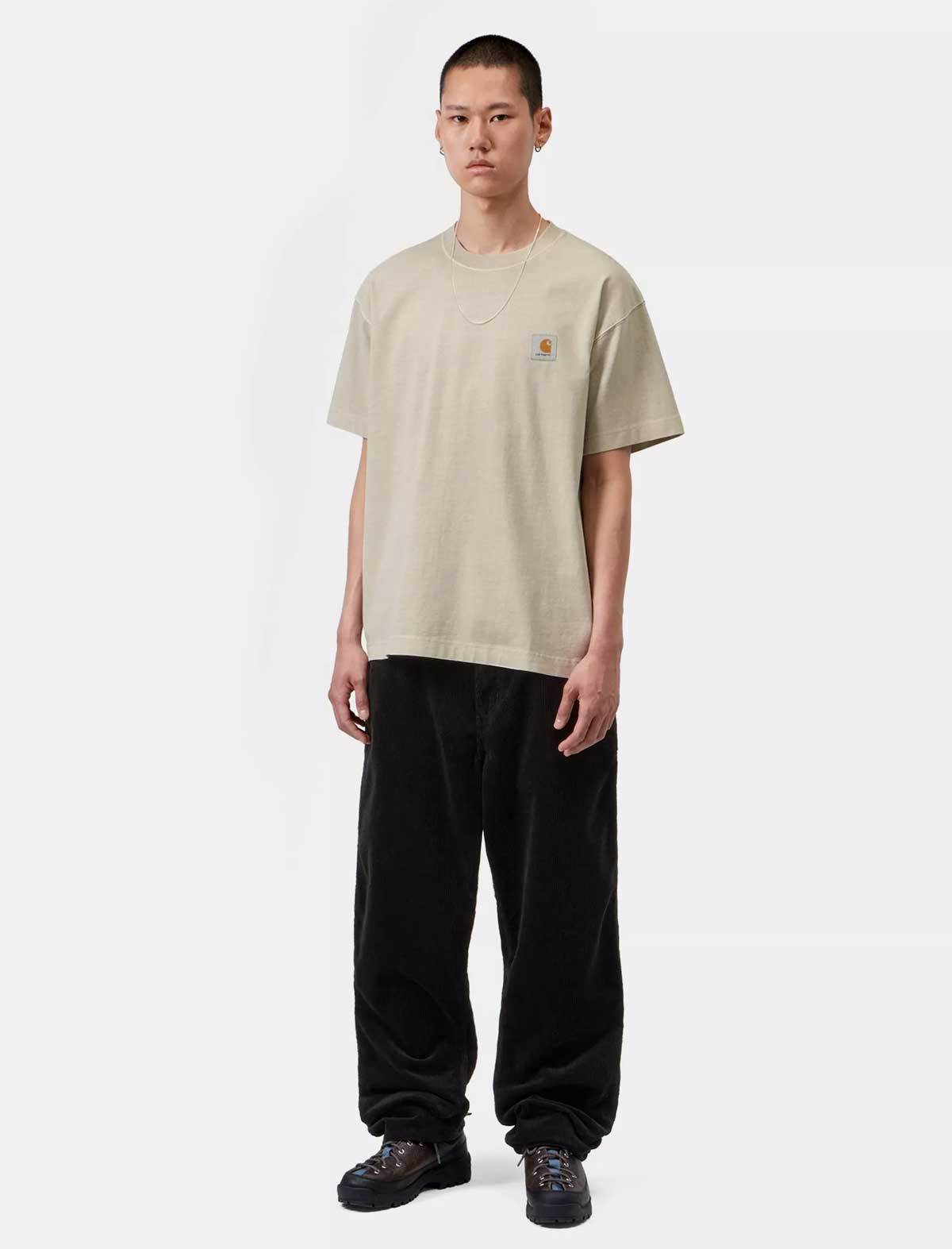 A model stands against a light gray studio background wearing a beige Carhartt t-shirt with the brand's signature chest logo, paired with black corduroy pants and black shoes.