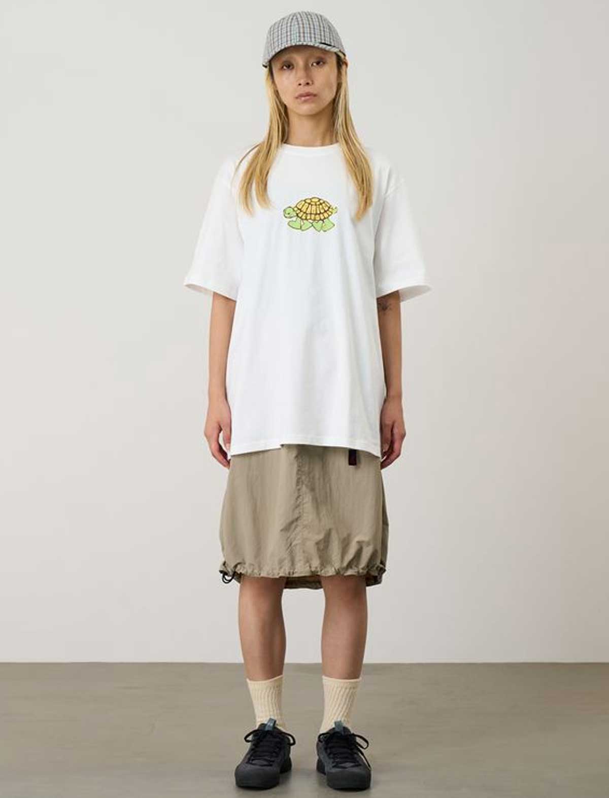Person wearing a white Gramicci Turtle t-shirt with a graphic design, beige skirt, and black shoes on a plain background