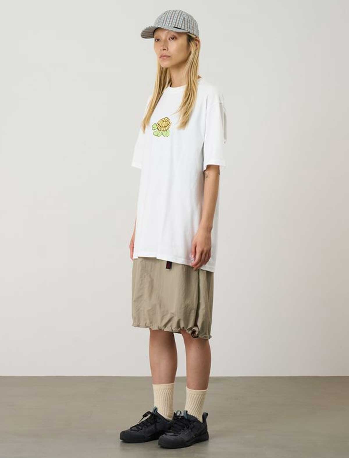 Person wearing a white t-shirt with a graphic design, beige shorts, and black shoes on a plain background