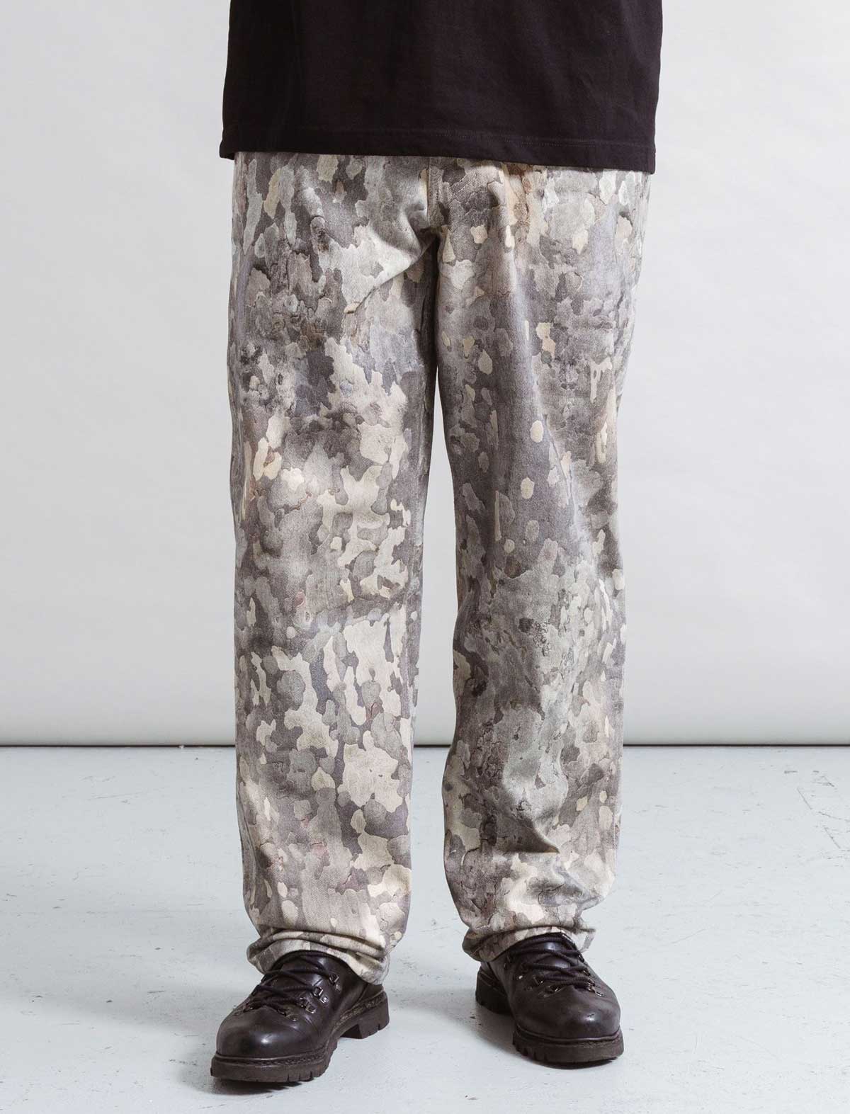 Person wearing Heresy Bark Jeans camouflage pants and black shoes on a white background