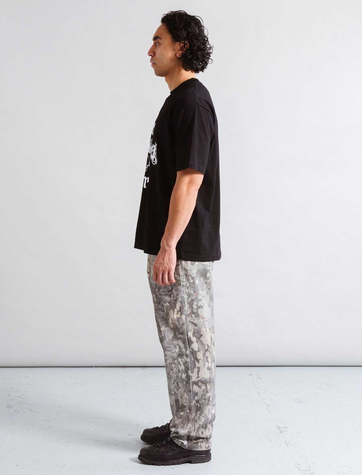 Person wearing a black t-shirt and camouflage pants standing against a white background