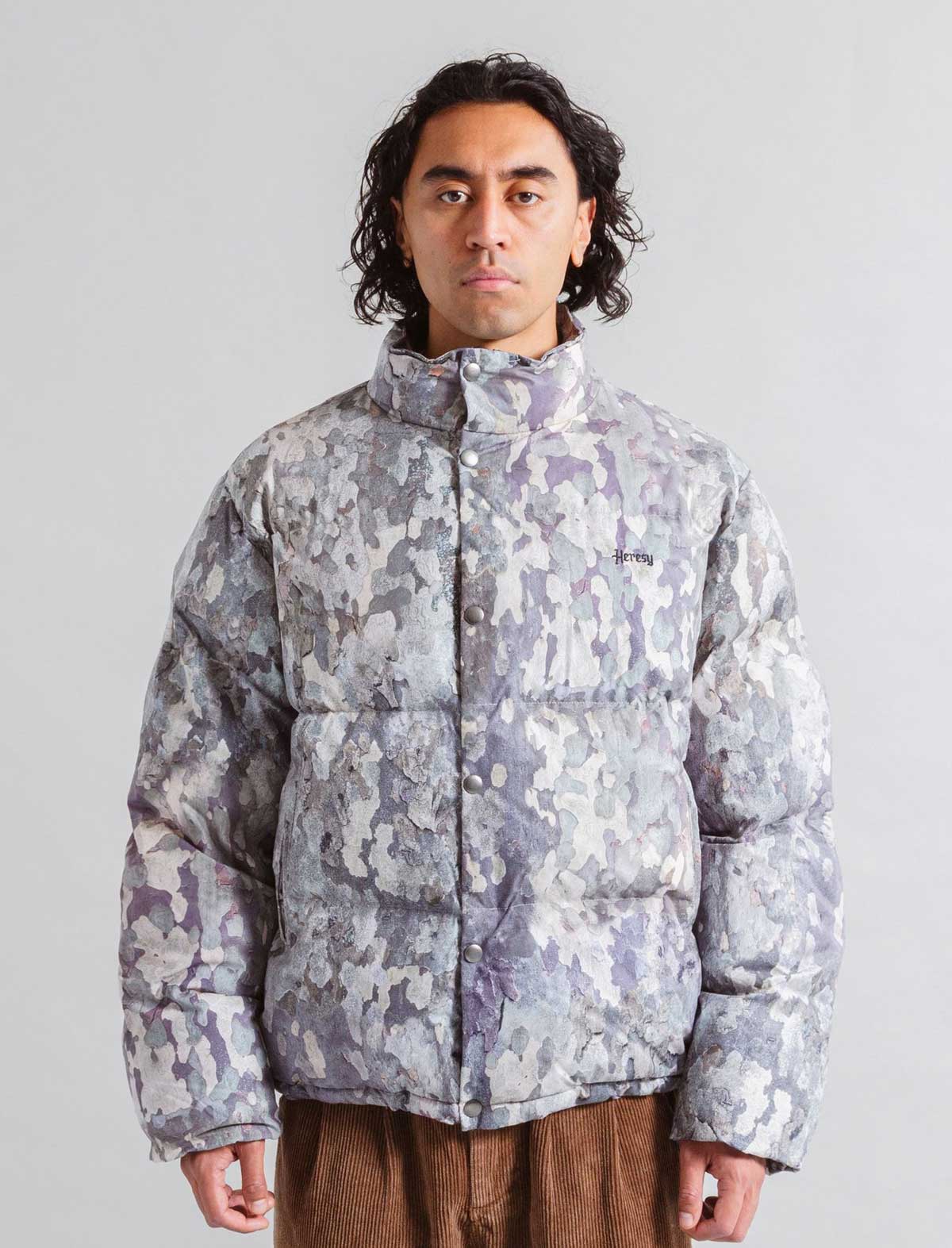 Person wearing a Heresy camouflage-patterned puffer jacket on a plain background