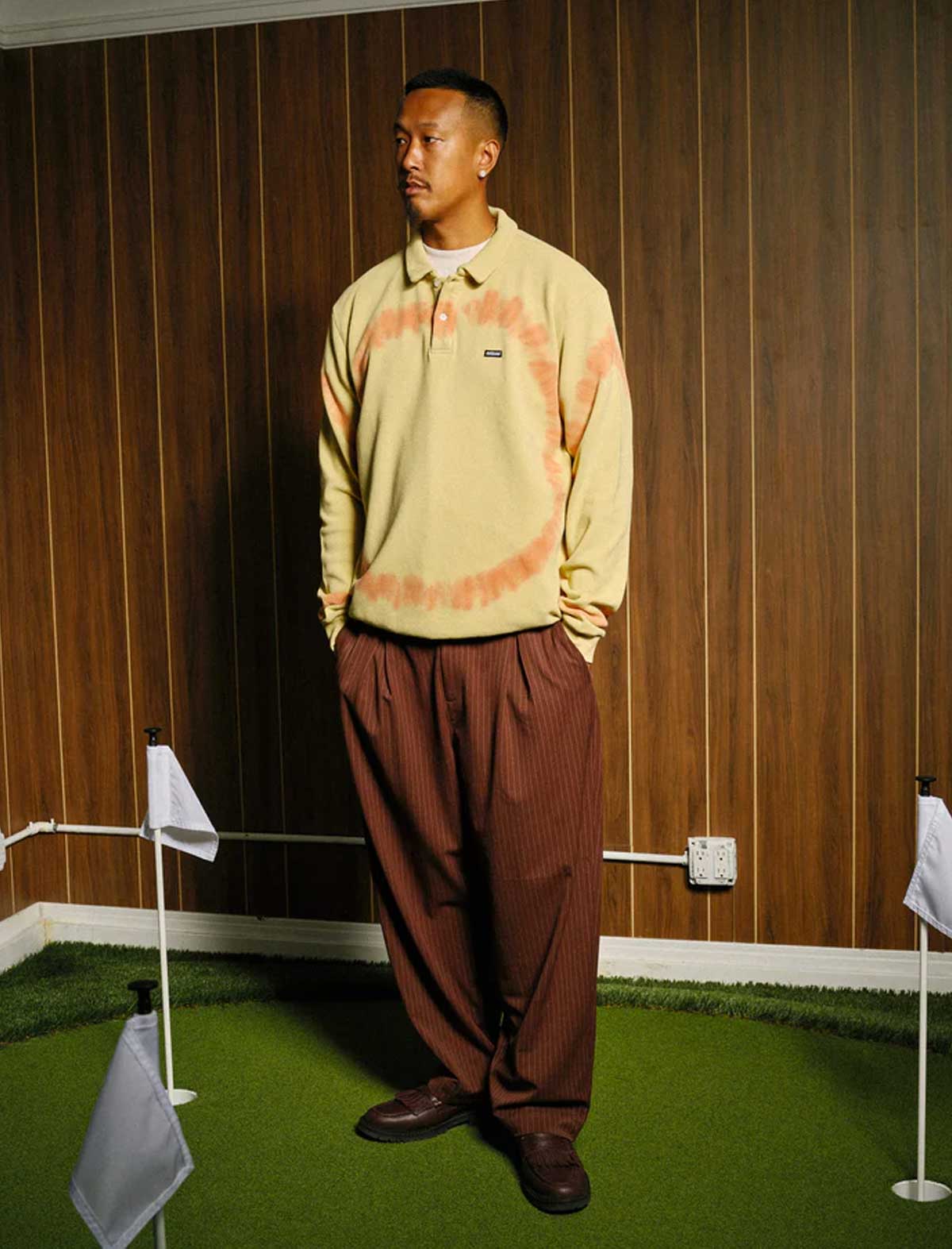 Man wearing a metalwood beige tie-dye sweater and brown pants standing in front of a wooden paneled wall.