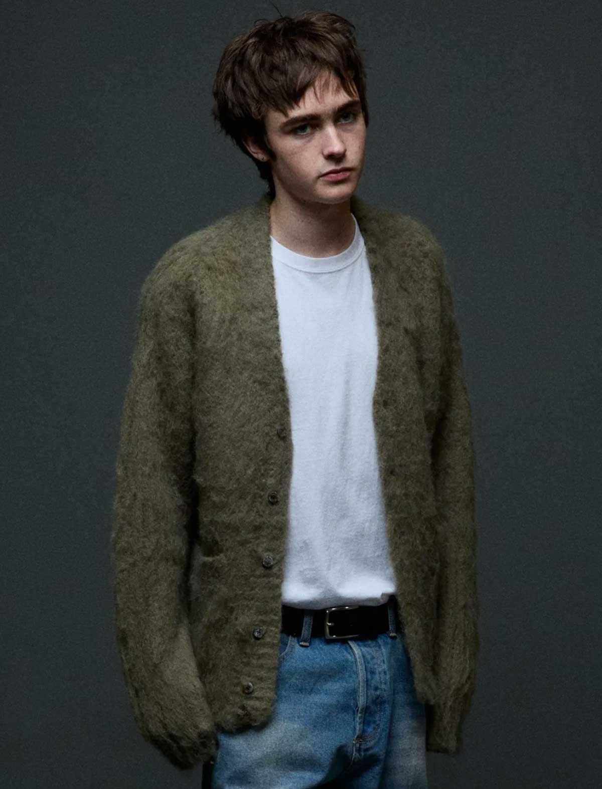 A young man wearing a fuzzy olive green cardigan over a white t-shirt and blue jeans, standing against a dark gray background.